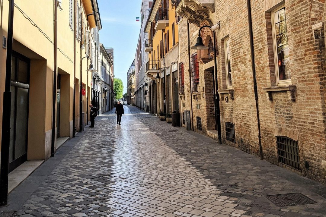 Narrow cobblestone street lined with old buildings, shopfronts, and a few people walking under clear skies.